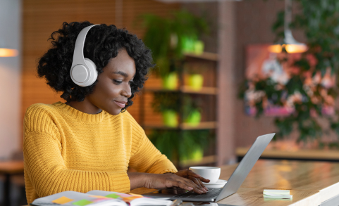 Femme portant un pull jaune et un casque audio blanc, travaillant sur un ordinateur portable dans un environnement chaleureux et lumineux avec des plantes en arrière-plan