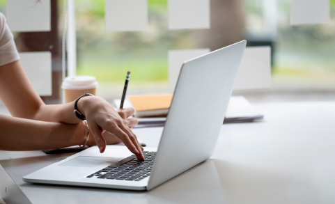 Main d'une personne utilisant un ordinateur portable sur un bureau, tenant un crayon dans l'autre main, avec une tasse de café et des cahiers en arrière-plan dans un cadre lumineux